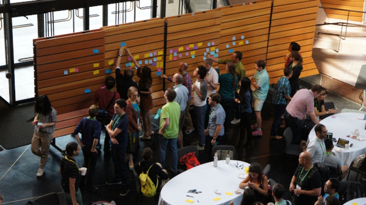 A crowd of about 20 people gathers in front of a board with a bunch of post it notes, as other participants sit at nearby tables in discussion.