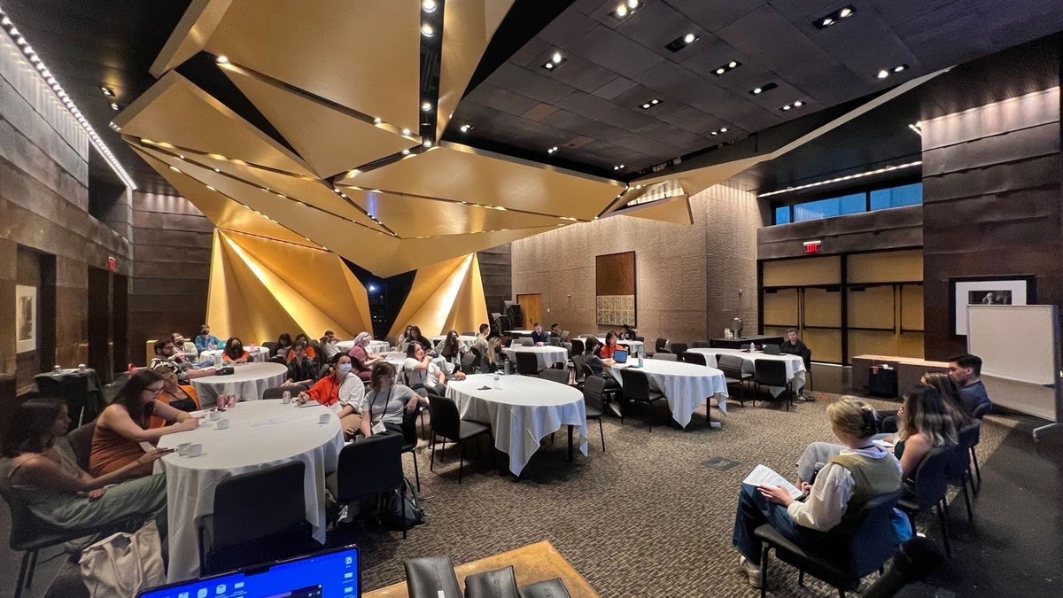 A Source-con session from 2024 in a conference space featuring round tables of early-career journalists listening to a panel at the front of the room.