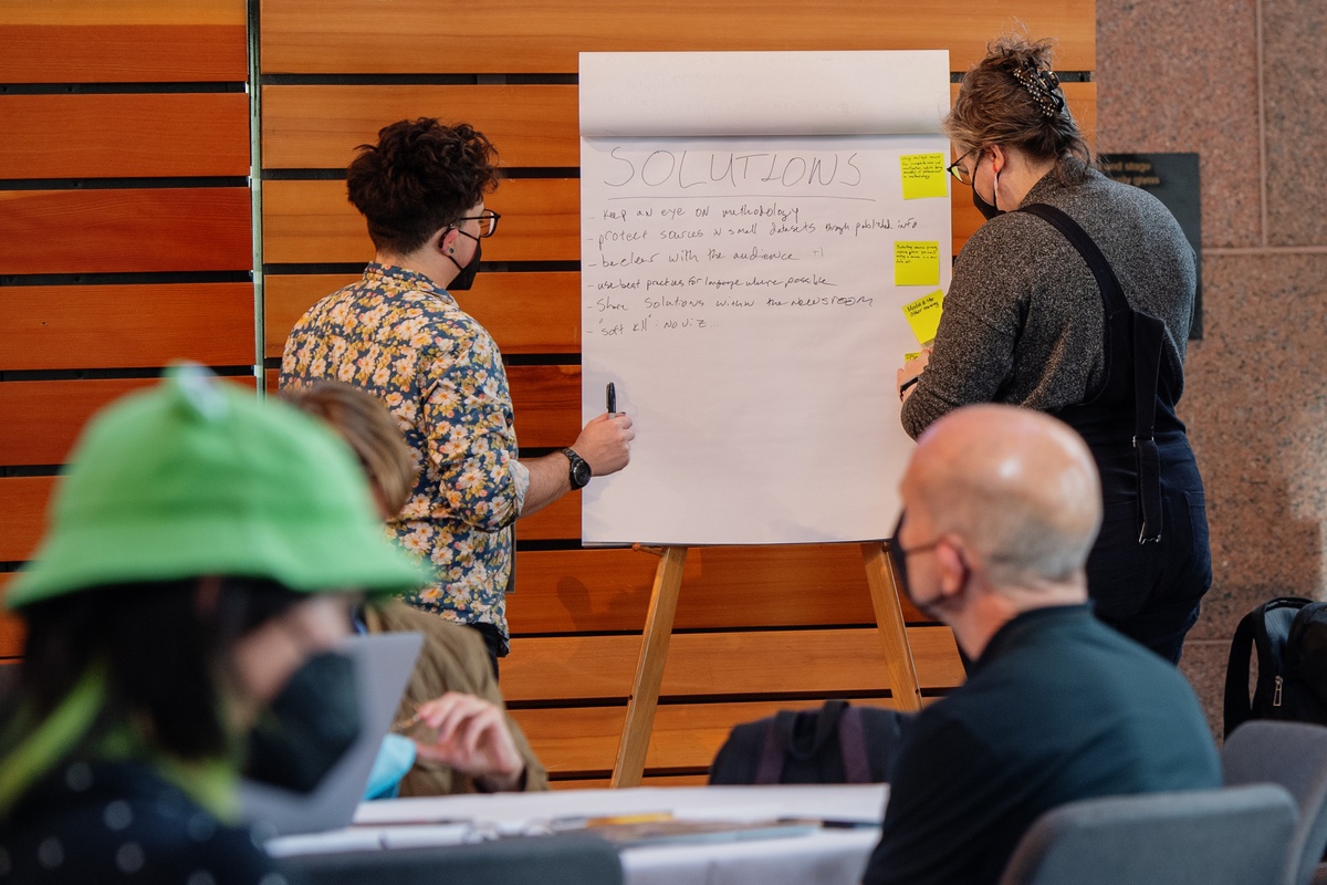 Session facilitators write notes on a big easel pad as workshop participants sit at tables during a workshop at SRCCON 2023.