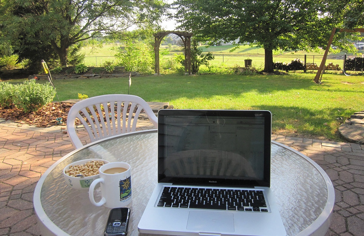 A laptop, phone, coffee, and breakfast cereal on a table looking out over a garden in the sun