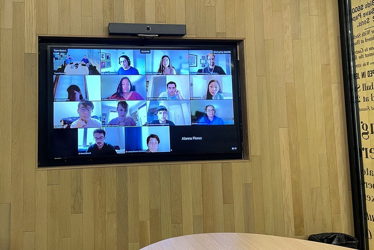 A TV screen on the wall of a meeting room, showing video thumbnails of participants in a Zoom meeting.