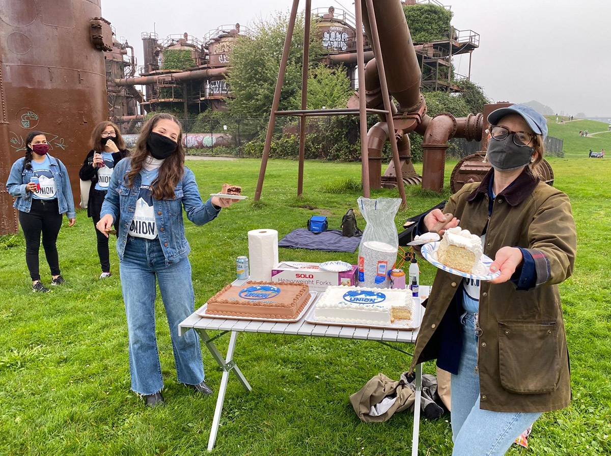 A woman wearing a cloth mask holds out a plate with a slice of cake on it, as other people gather around a picnic table in a park